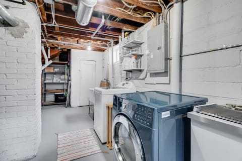 A clean basement laundry room featuring a washing machine, utility sink, and shelves with cleaning supplies.