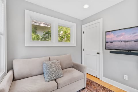 A light grey room featuring a sofa, windows with greenery, and a mounted TV displaying sailboats.