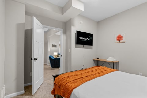 A bedroom with an orange throw blanket, a mounted TV, and a doorway leading to another room.