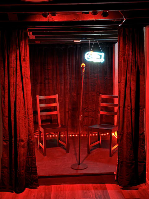 A small performance stage with a microphone and two chairs, lit in red with a neon sign overhead.