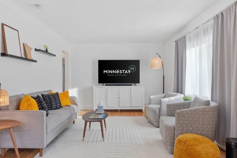 Cozy living room with a gray sofa, yellow accents, and a TV displaying the MINNēSTAY logo.