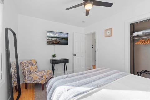 A minimalist bedroom featuring a bed, floral chair, mounted TV, full-length mirror, and organized closet.