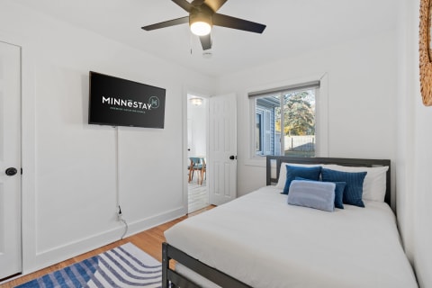 A bright and airy bedroom with a bed, blue decorative pillows, a flat-screen TV, and a window view.