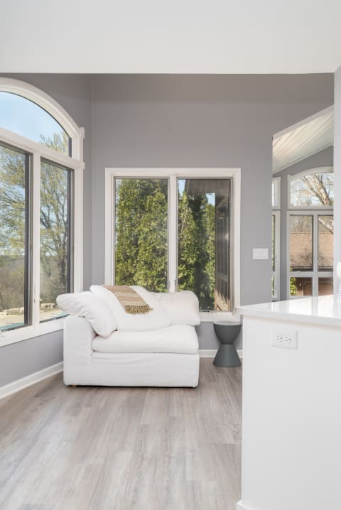 Bright corner of a room with large windows, a white loveseat, and a light wood floor.