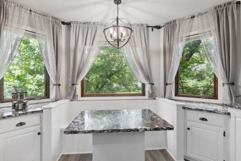 A small kitchen area featuring a marble table and large windows with curtains, showcasing greenery outside.