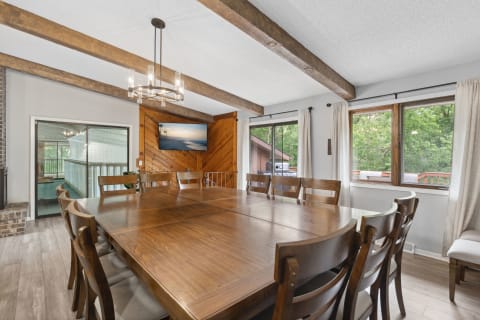 A dining room featuring a large wooden table, modern chandelier, and windows overlooking greenery.