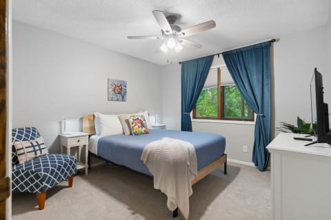A well-decorated bedroom featuring a bed, armchair, window with greenery outside, and a television.