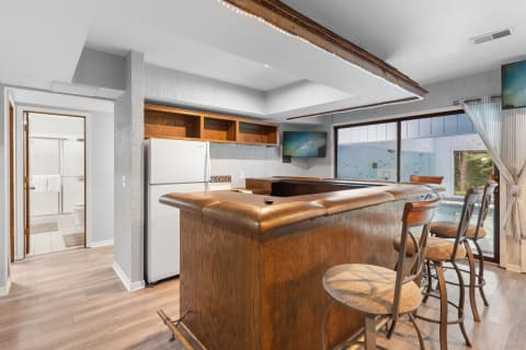 Interior view of a kitchen bar with wooden counter, stools, a refrigerator, and a bathroom door.