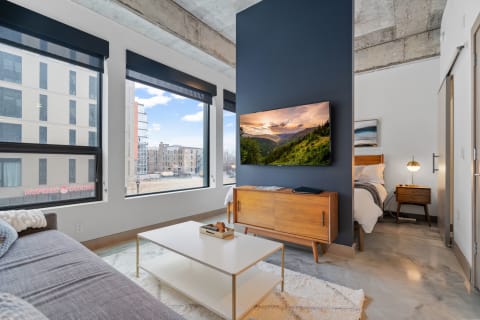 Modern living room with large windows, a TV, and a cozy bed in the background.