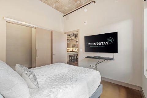Stylish modern bedroom with a white bed, sliding closet door, and a mounted television displaying the MINNESTAY logo.