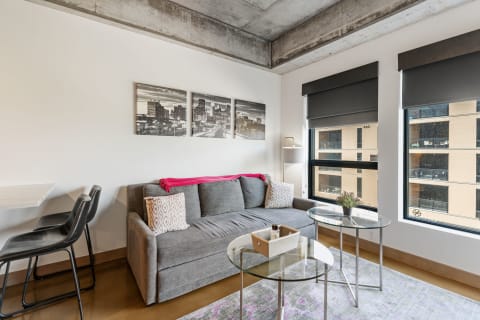 A contemporary living room featuring a gray sofa with cushions, glass coffee tables, cityscape art, and large windows.