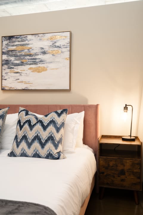 A cozy bedroom with a pink bed, decorative pillows, and a modern nightstand with a lamp.
