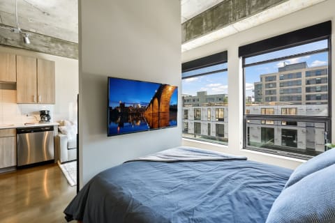 Modern bedroom with a wall-mounted TV displaying a city skyline, kitchenette and window views.