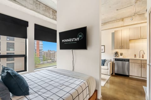 Cozy studio apartment with a bed and kitchen area, large windows showing an urban view.