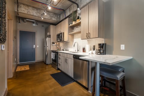 A modern kitchen with light wood cabinetry and a quartz countertop, showcasing stainless steel appliances and a warm concrete floor.