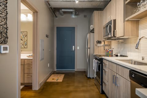 A view of a modern kitchen and entryway with light wood cabinets and a blue front door.