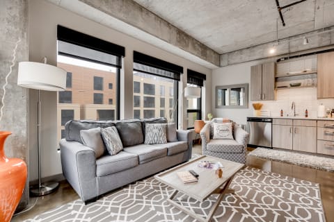A modern living room featuring a grey sofa, armchair, coffee table, and a compact kitchen in the background.