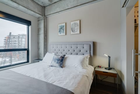 Modern bedroom with a tufted headboard, bedside table, and urban views through a large window.