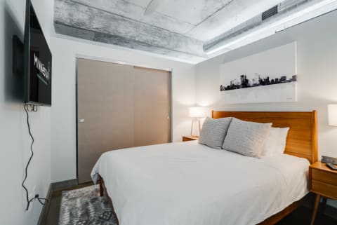 A minimalist bedroom with a white bed, wooden headboard, and a skyline artwork.