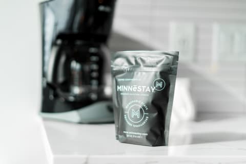 A black coffee packet labeled MINNĒSTAY sits on a marble countertop with a coffee maker in the background.
