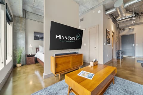 Interior view of a modern living room with MINNēSTAY branding, featuring wooden furniture, a large TV, and a cozy atmosphere.