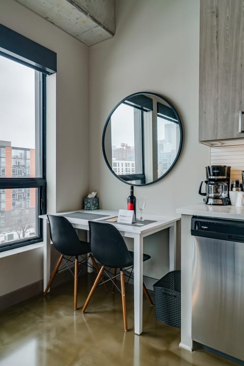 A small dining area in a modern kitchen features a white table, black chairs, a wine bottle, and large round mirror reflecting the city outside.