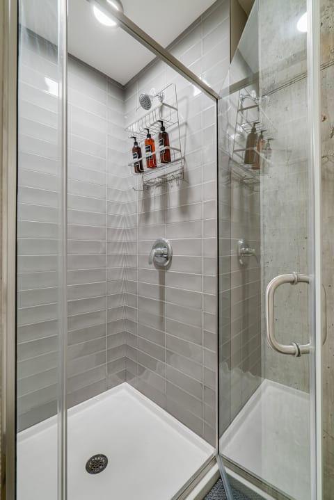 A modern shower with gray tiles and a chrome showerhead, featuring organized bottles on a wire shelf.