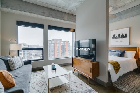 A stylish studio apartment featuring a sofa, coffee table, TV, and a neatly made bed with city views.
