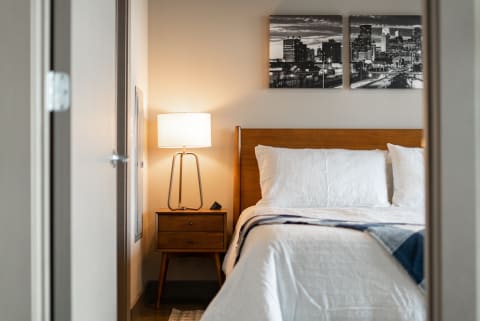 A modern bedroom featuring white bedding, a wooden bedside table, and black-and-white city skyline artwork.