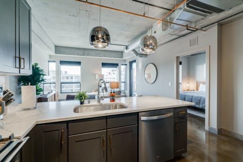 A modern kitchen with a white quartz countertop and dark cabinetry, opening to a living area with large windows.