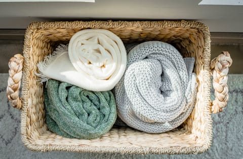A woven basket with three rolled blankets: one white, one green, and one grey.