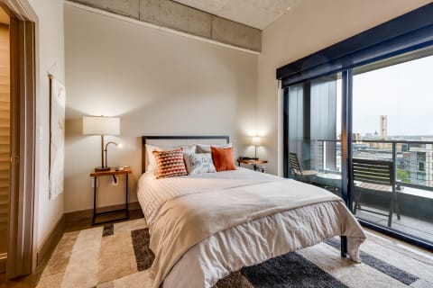 A modern bedroom featuring a bed with decorative pillows, a nightstand with a lamp, and a balcony view.