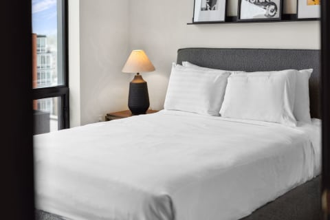 A modern bedroom featuring a white bed with pillows, a black lamp, and framed artwork above.