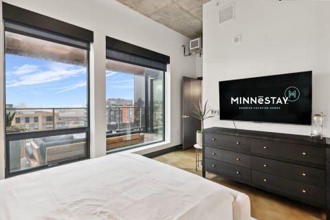 Contemporary bedroom with large windows and city view, featuring a bed, dresser, and TV.