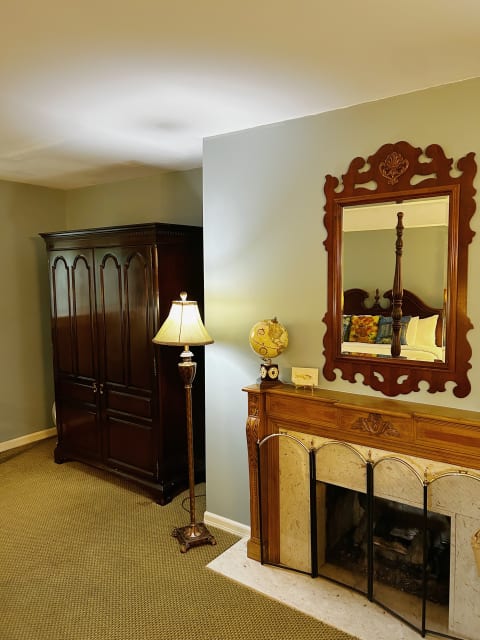A warm and inviting room featuring a wooden armoire, a floor lamp, an ornate mirror above a fireplace mantel, and decorative items.