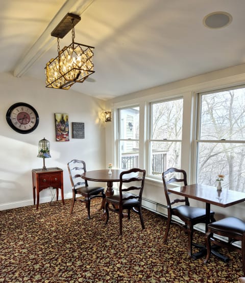 A cozy dining room with wooden furniture, a decorative chandelier, and large windows showcasing a view of trees.