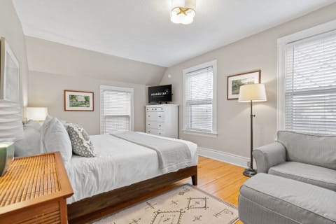 Cozy bedroom featuring a bed, gray couch, lamp, and television with natural light from windows.