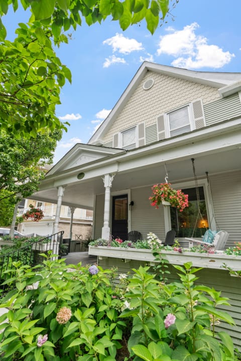 A light gray house with a flower-adorned porch under a blue sky with clouds.