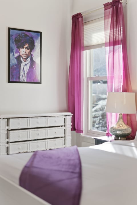 A cozy bedroom with purple accents, featuring a framed artistic portrait, stylish curtains, and a vintage dresser.