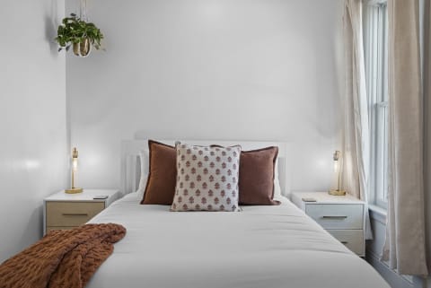 A serene bedroom with a made bed, decorative pillows, a cozy throw, a nightstand with a lamp, and a hanging plant.