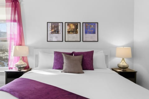 A stylish bedroom featuring white linens, purple accents, framed musical posters, and lamps.