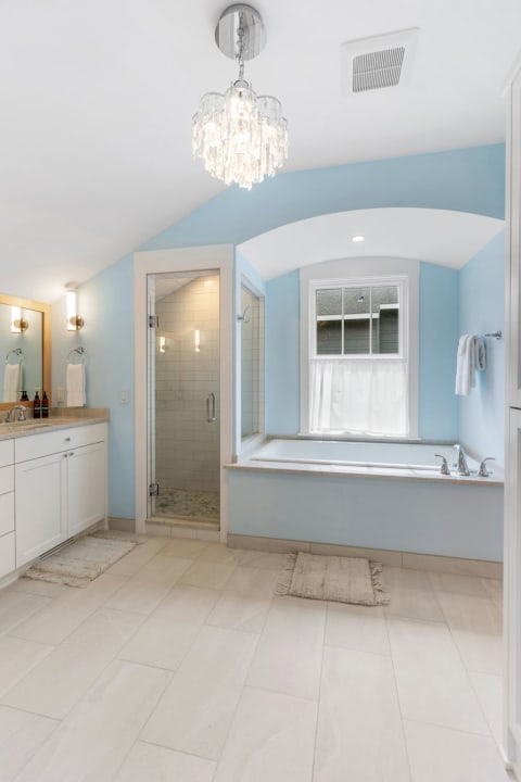 Elegant bathroom with light blue walls, crystal chandelier, glass shower, and soaking tub.