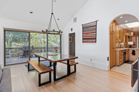 A dining area featuring a glass table with wooden benches, a chandelier, and an open kitchen.