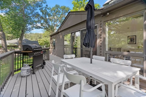 A spacious deck featuring a grill and a white dining table with chairs, set against a backdrop of trees and a blue sky.