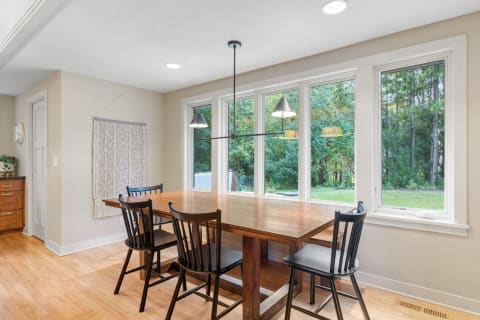 Bright dining area with a wooden table, black chairs, and large windows overlooking greenery.
