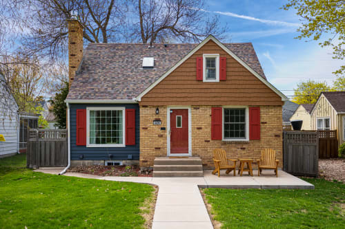 Charming brick cottage with red shutters and welcoming front patio featuring wooden chairs for relaxing outdoors.