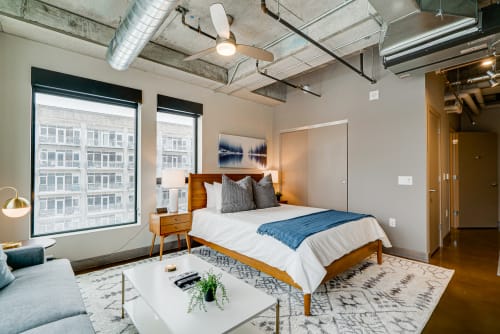 Relax in your stylish urban loft featuring a plush bed, exposed ceilings, and stunning city views through oversized windows.
