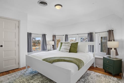 Sink into crisp white linens in this bright corner bedroom, where morning light streams through multiple windows and nightstands holds bedside essentials.